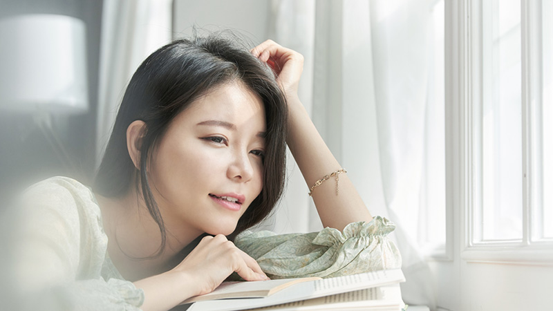 窓際で眠そうな表情で本を開く女性